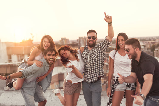 Sechs fröhliche Männer und Frauen im Alter von ca. 22 Jahre tanzen auf einer Terrasse. Im Hintergrund strahlt die Sonne.