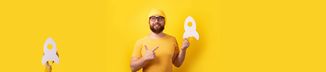 Eine männliche Person (frontal zeigend), um die 30 Jahre alt, mit gelbem T-Shirt und gelber Mütze, hält eine Papier-Rakete nach oben zeigend in der Hand. Auch der Hintergrund ist gelb.