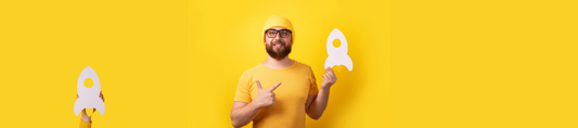 Eine männliche Person (frontal zeigend), um die 30 Jahre alt, mit gelbem T-Shirt und gelber Mütze, hält eine Papier-Rakete nach oben zeigend in der Hand. Auch der Hintergrund ist gelb.