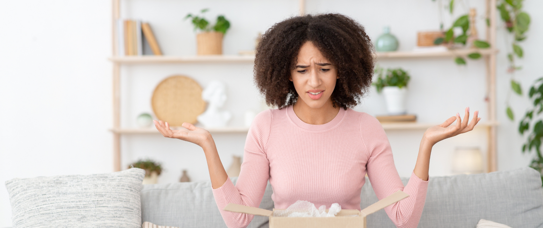 Eine weibliche afroamerikanische Person sitzt auf einem Sofa und hat auf ihren Beinen ein geöffnetes Paket, links und rechts sind beide Arme unglaubwürdig in der Luft und schaut auch fraglich zum Paket. 