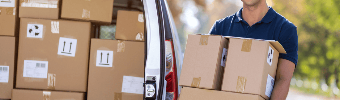 Ein Lieferbote (Mann) steht neben seinem Lieferwagen (hinten) und hat mehrere Pakete in seinen Händen. 