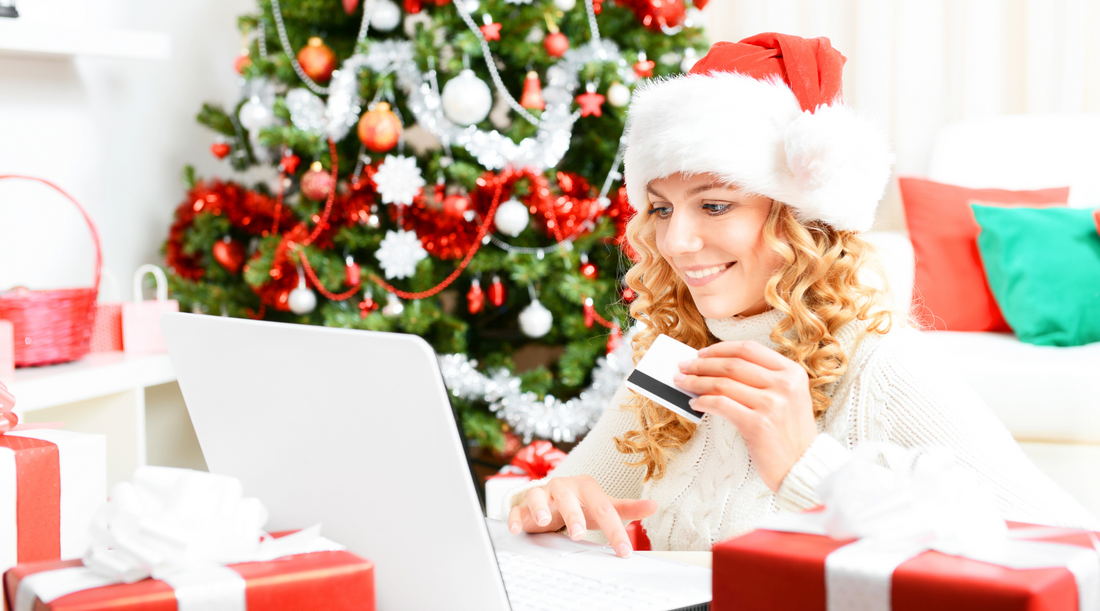 Frau kauft online Weihnachtsgeschenke mit Kreditkarte am Laptop vor festlich geschmücktem Weihnachtsbaum.
