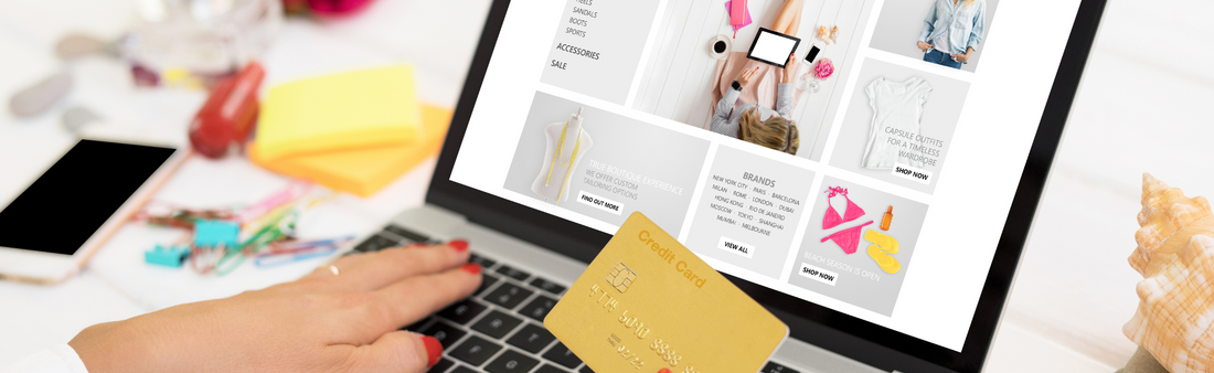 Eine nicht zu sehende Person (nur die Hände) an einem Schreibtisch, an einem geöffneten Laptop beim Online-Shoppen.