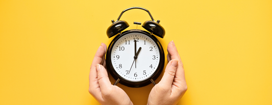 Zwei Hände umgreifen schützend einen älteren, mechanischen Uhrwecker vor einem dunkelgelben Hintergrund, symbolisch für die Zeitumstellung und Chronobiologie.