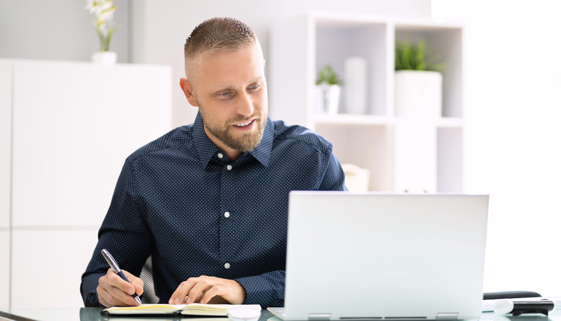 Mann arbeitet konzentriert am Laptop im Büro und macht Notizen – produktives Arbeiten im modernen Arbeitsumfeld.