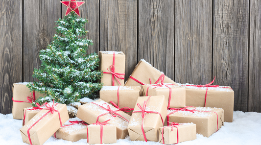 Kleiner Weihnachtsbaum im Schnee, umgeben von vielen braunen Geschenkpaketen mit roten Bändern vor rustikaler Holzwand.