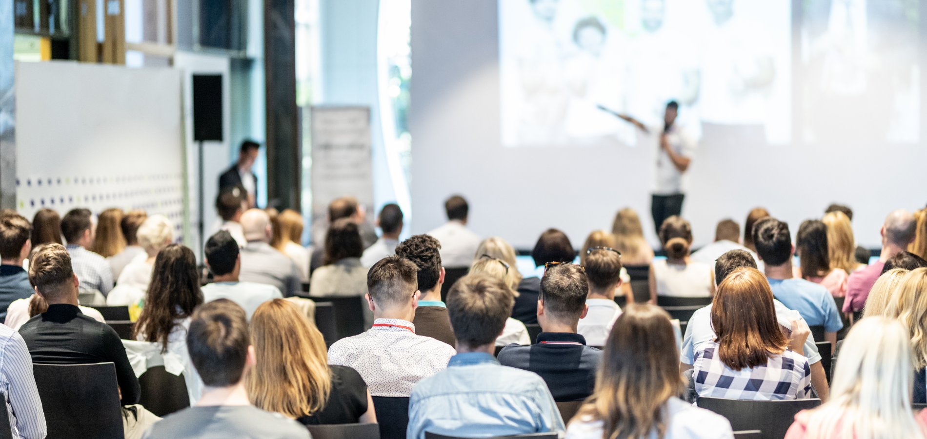 Seminarveranstaltung: Ca. 40 Personen schauen Richtung Bühne und hören einem männlichen Vortragsredner auf der Bühne zu.