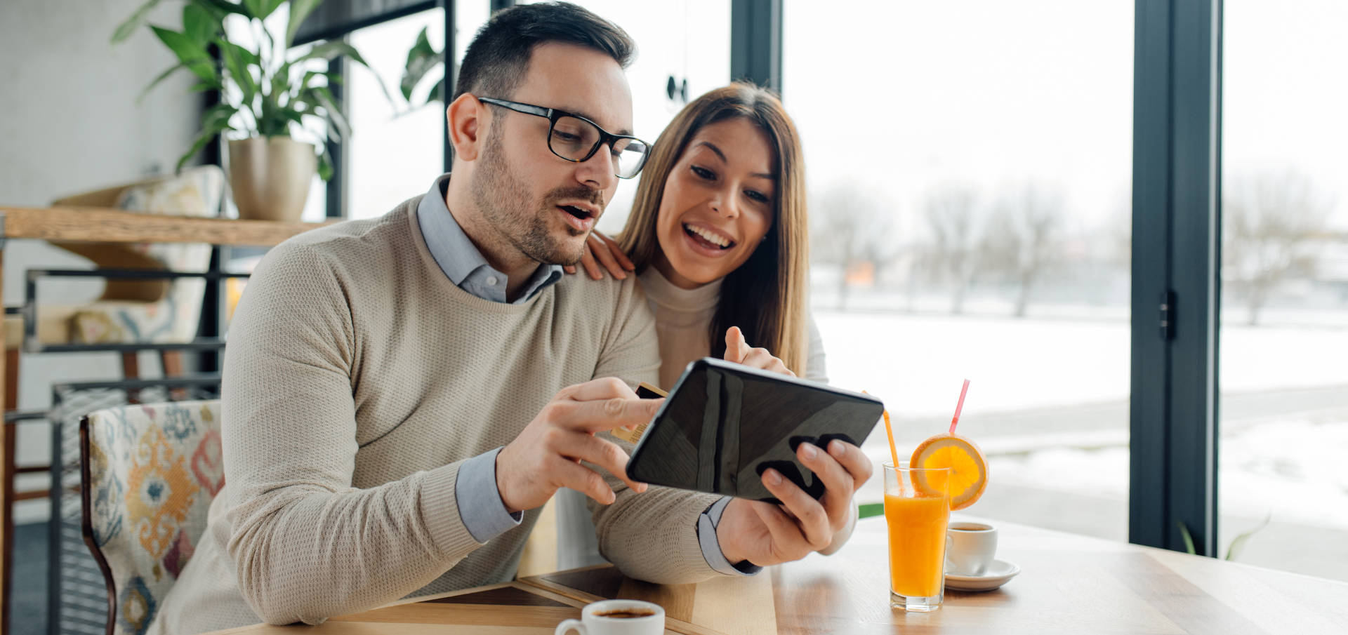 Ein junger Mann und eine Frau sitzen geminsam an einem Tisch sowie schauen sie lachend auf ein Tablet und shoppen 