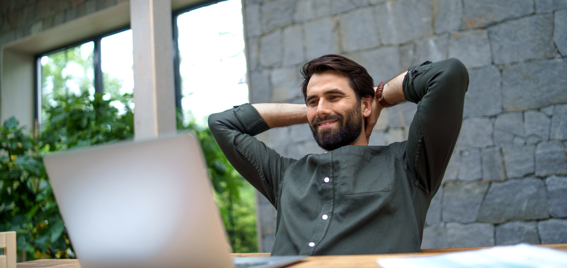 Entspannter Mann sitzt lächelnd vor Laptop im modernen Büro mit Naturblick. Abgesichert und Zufriedenheit Konzept