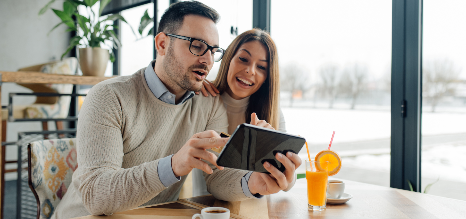 Lächelndes Paar im Café nutzt Tablet beim Online-Shopping, mit Kaffee und Orangensaft auf dem Tisch