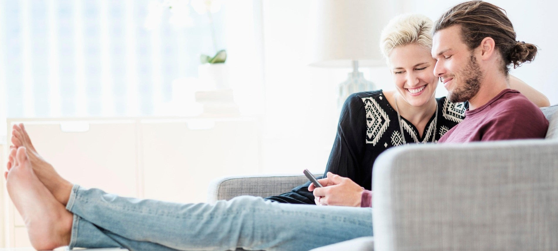 Lächelndes Paar sitzt entspannt auf dem Sofa und schaut gemeinsam auf ein Smartphone, glücklicher Moment im Alltag.