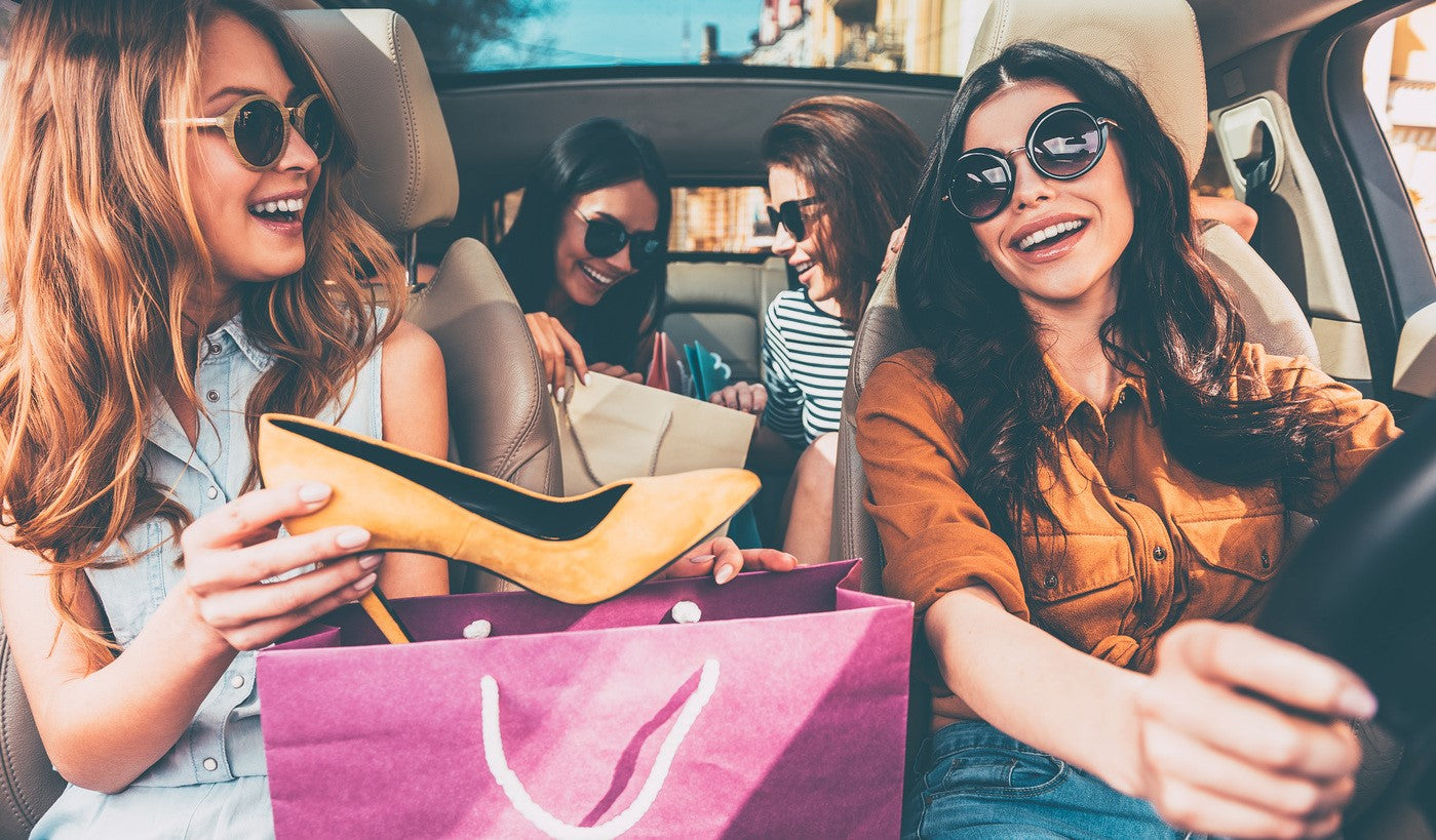 Vier junge Frauen lachen im Auto nach dem Shopping, zeigen Einkäufe und genießen gemeinsam Lifestyle und Shoppingfreude.